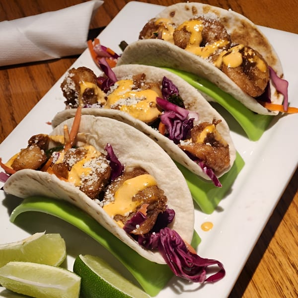 Three SamB's American-style tacos with fried shrimp, red cabbage, sauce, and lime wedges.
