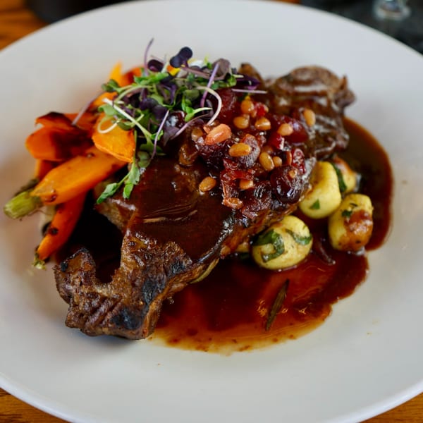 Braised short rib with roasted carrots, gnocchi, and a rich sauce, garnished with microgreens.