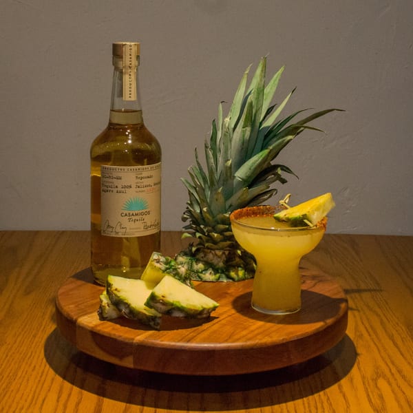 Casamigos tequila bottle, whole pineapple, pineapple slices, and a pineapple margarita on a wooden tray.