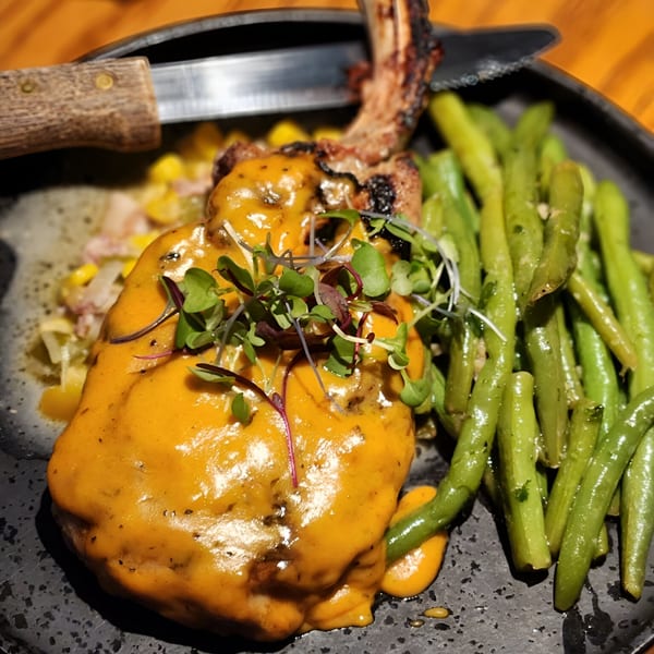 Pork chop with orange sauce, microgreens, green beans, and corn on a black plate.