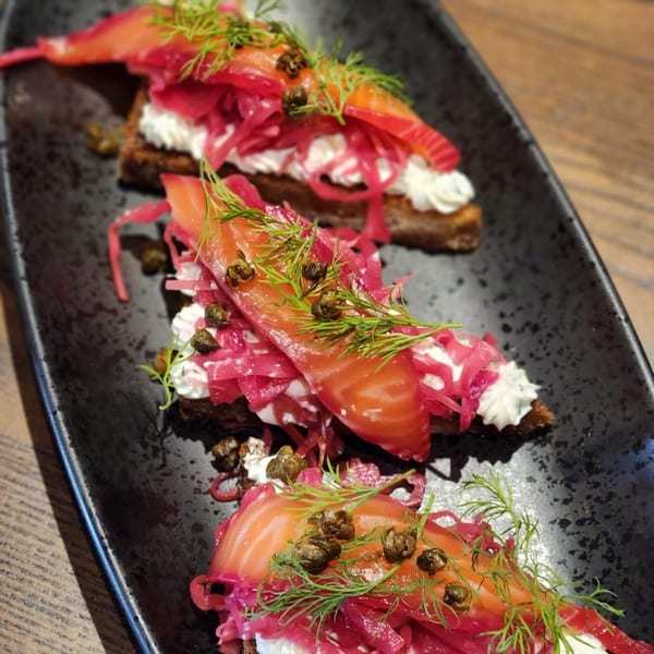Three salmon and cream cheese toasts with pickled red onions, capers, and dill on a black plate.