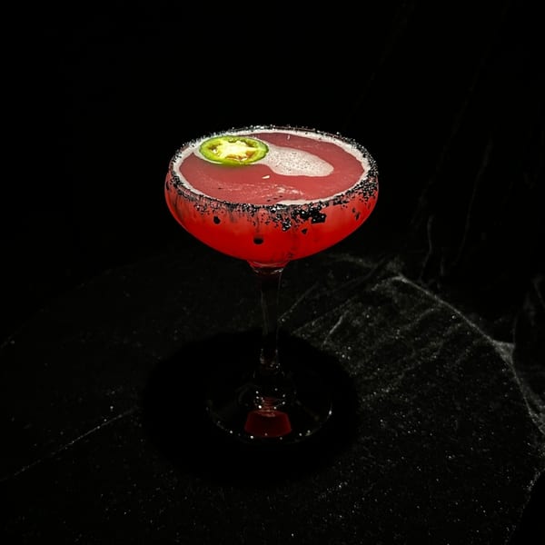 Red cocktail with a black sugar rim and a green jalapeño slice, against a dark background.