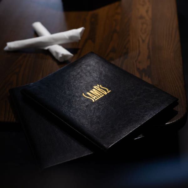 Two black SamB's menus with gold lettering and two rolled white napkins on a dark wooden table.