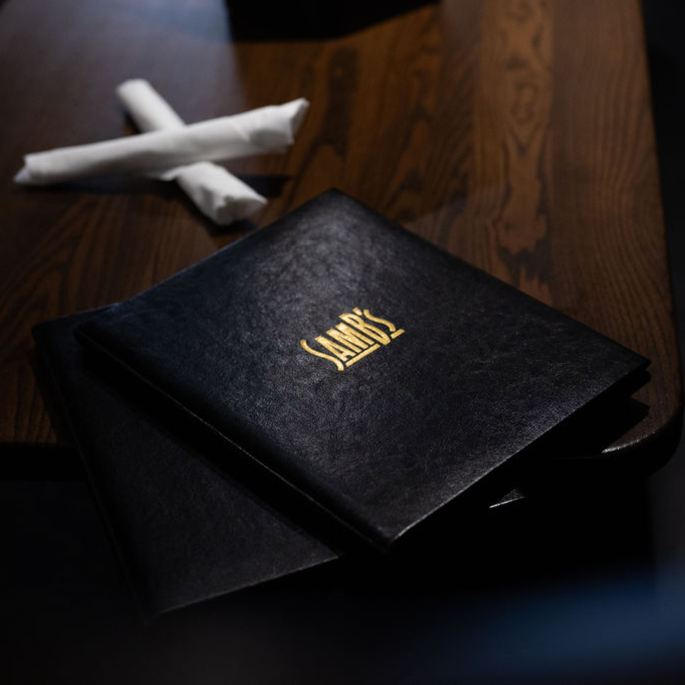 Two black SamB's menus with gold lettering and two rolled white napkins on a dark wooden table.