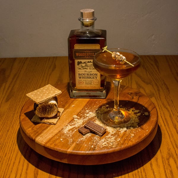 S'mores dessert with a bourbon cocktail and bottle of Woodinville Straight Bourbon Whiskey on a wooden board.