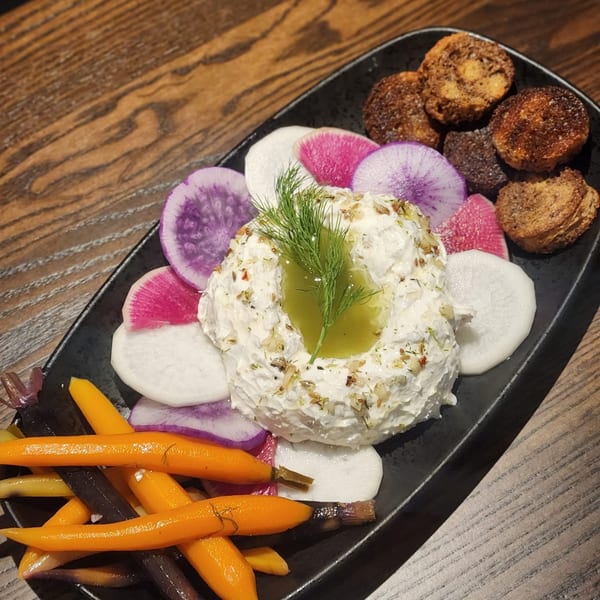 A black platter with a creamy dip, colorful radish slices, roasted carrots, and bread pieces.