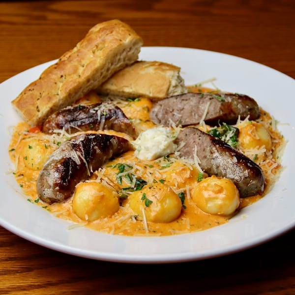 A white plate of gnocchi and sausage in a creamy sauce, topped with cheese and focaccia bread.