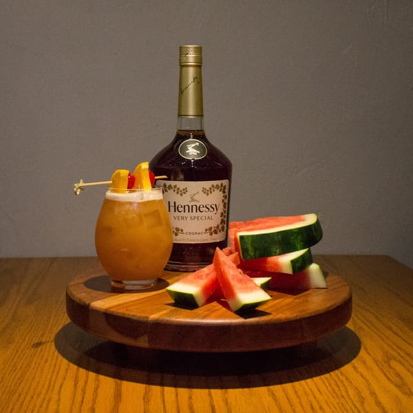A Hennessy bottle, a cocktail with fruit garnish, and watermelon slices on a wooden tray.
