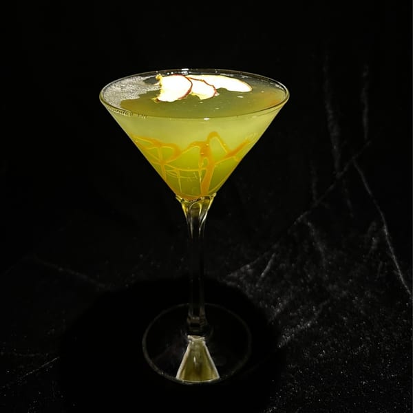 A green cocktail in a martini glass, garnished with apple slices and caramel drizzle.