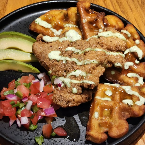 Chicken and waffles with avocado slices and pico de gallo on a black plate.